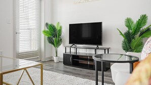 A modern living room with a flat-screen TV, two potted plants, a glass coffee table, and a white sofa. - Stylish Stay | Near Everything! (Whitestown)