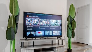 A flat-screen TV on a wooden TV stand with a YouTube Sports channel interface, flanked by large green plants. - Refined Comforts: Luxe Stay in Whitestown Indiana (Perry Township)