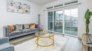 A modern living room with a grey sofa, a glass coffee table, and a large window with a view of buildings. - Refined Comforts: Luxe Stay in Whitestown Indiana (Perry Township)