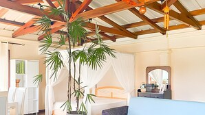 A spacious room with a high ceiling and exposed wooden beams, featuring a large potted plant, a blue sofa, a small wooden table, and a bed with white curtains.
