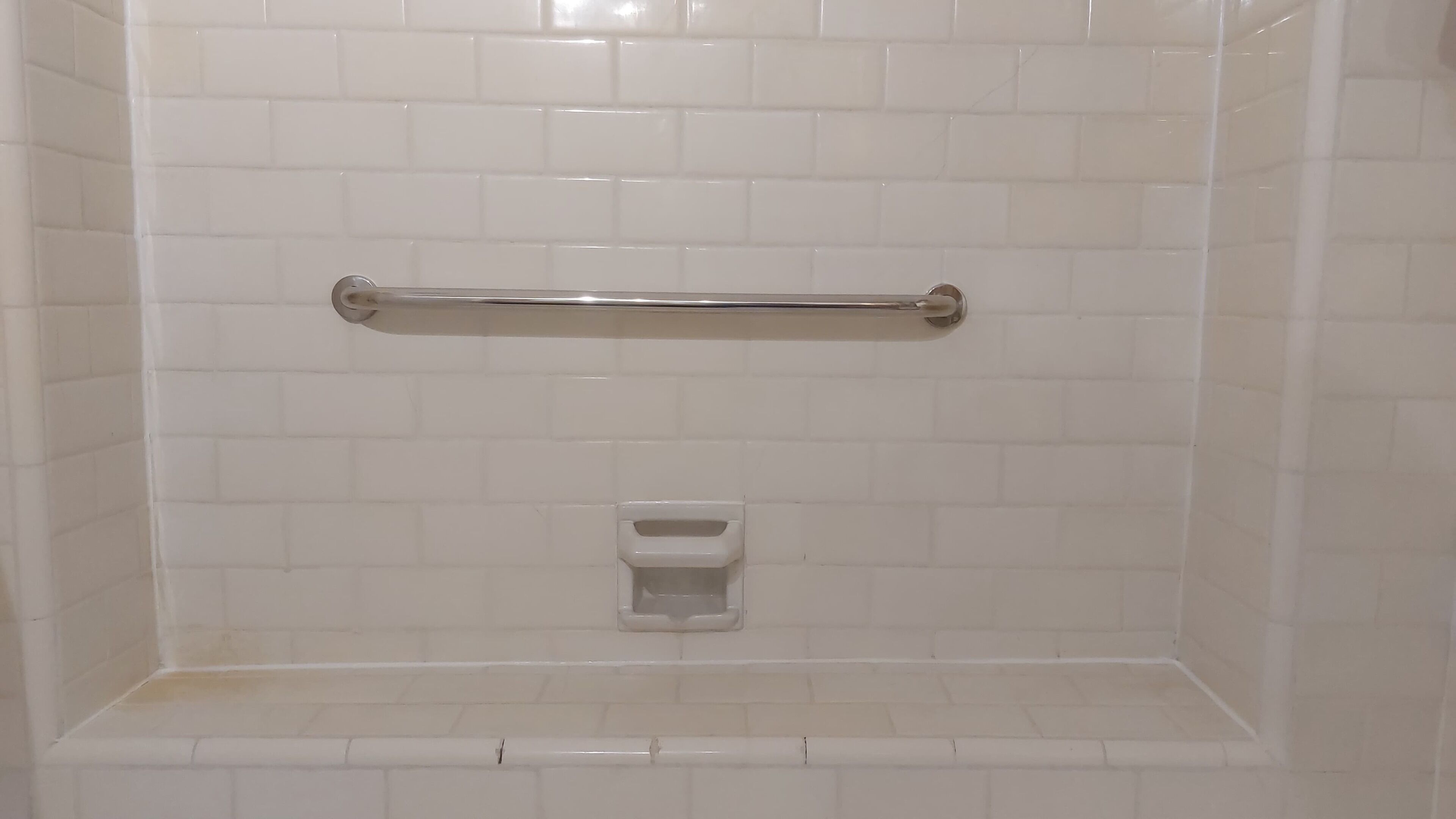Una ducha con paredes de azulejos blancos, una barra de apoyo metálica y un pequeño lavamanos pedestal blanco.