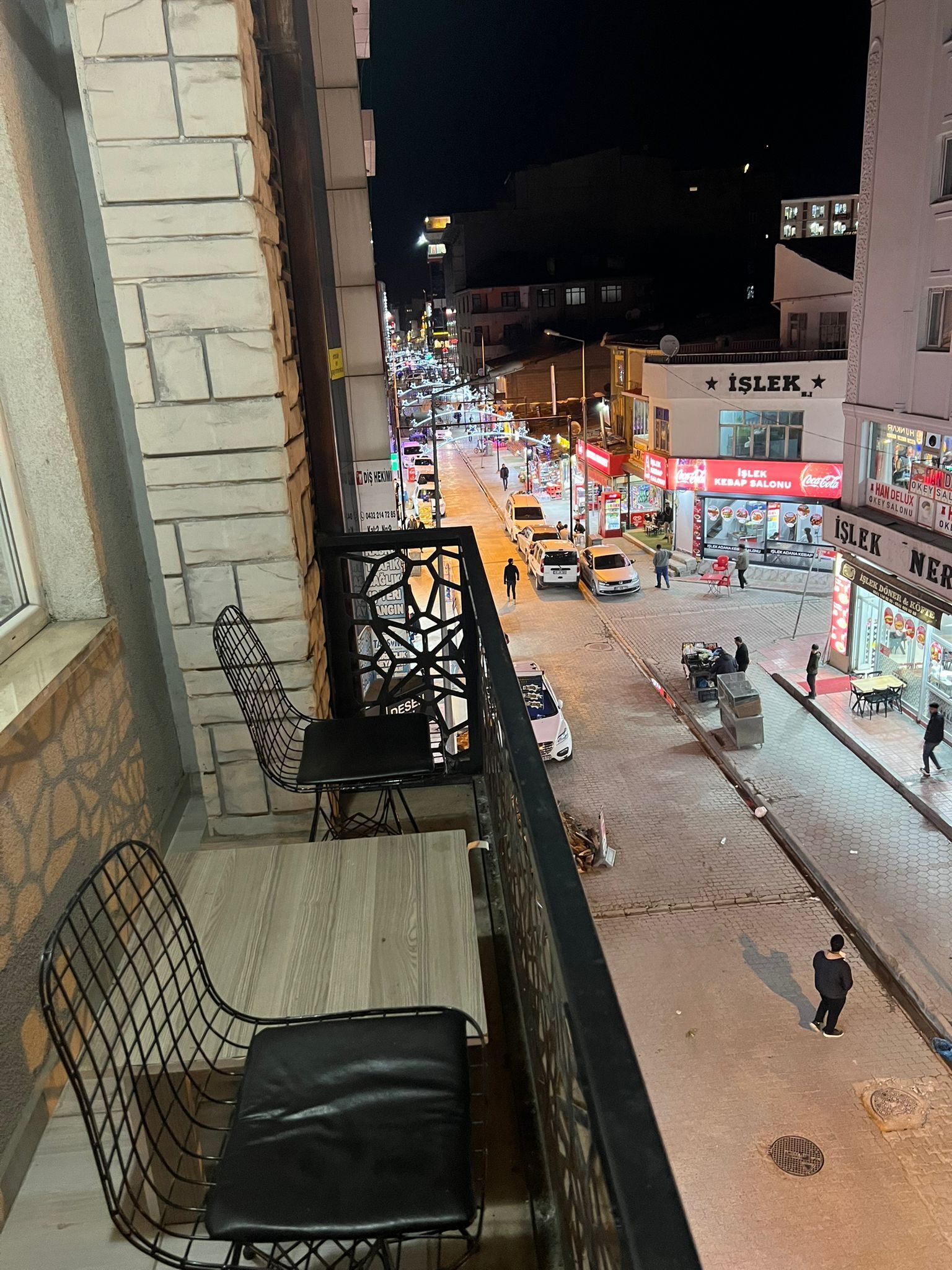 A balcony with two chairs and a table, overlooking a street with shops and cars.