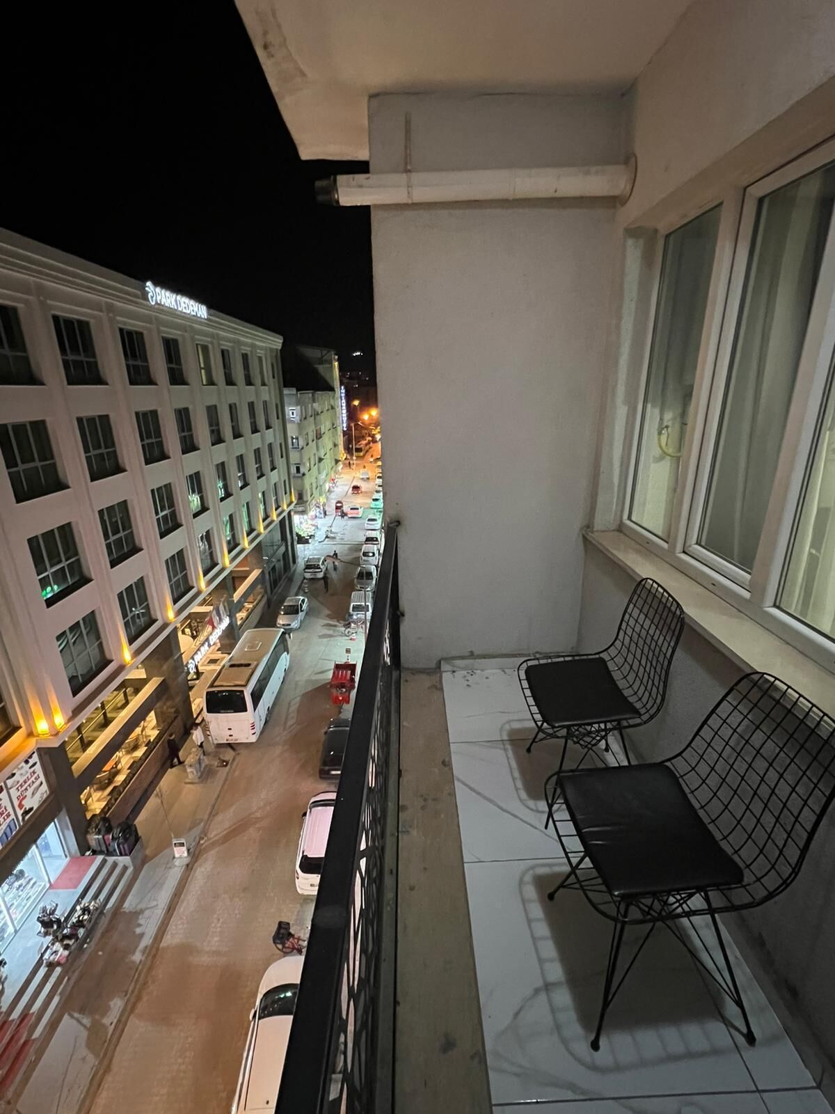 Uma varanda com duas cadeiras de metal preto, vista para a rua com carros estacionados e um edifício com letreiros iluminados.
