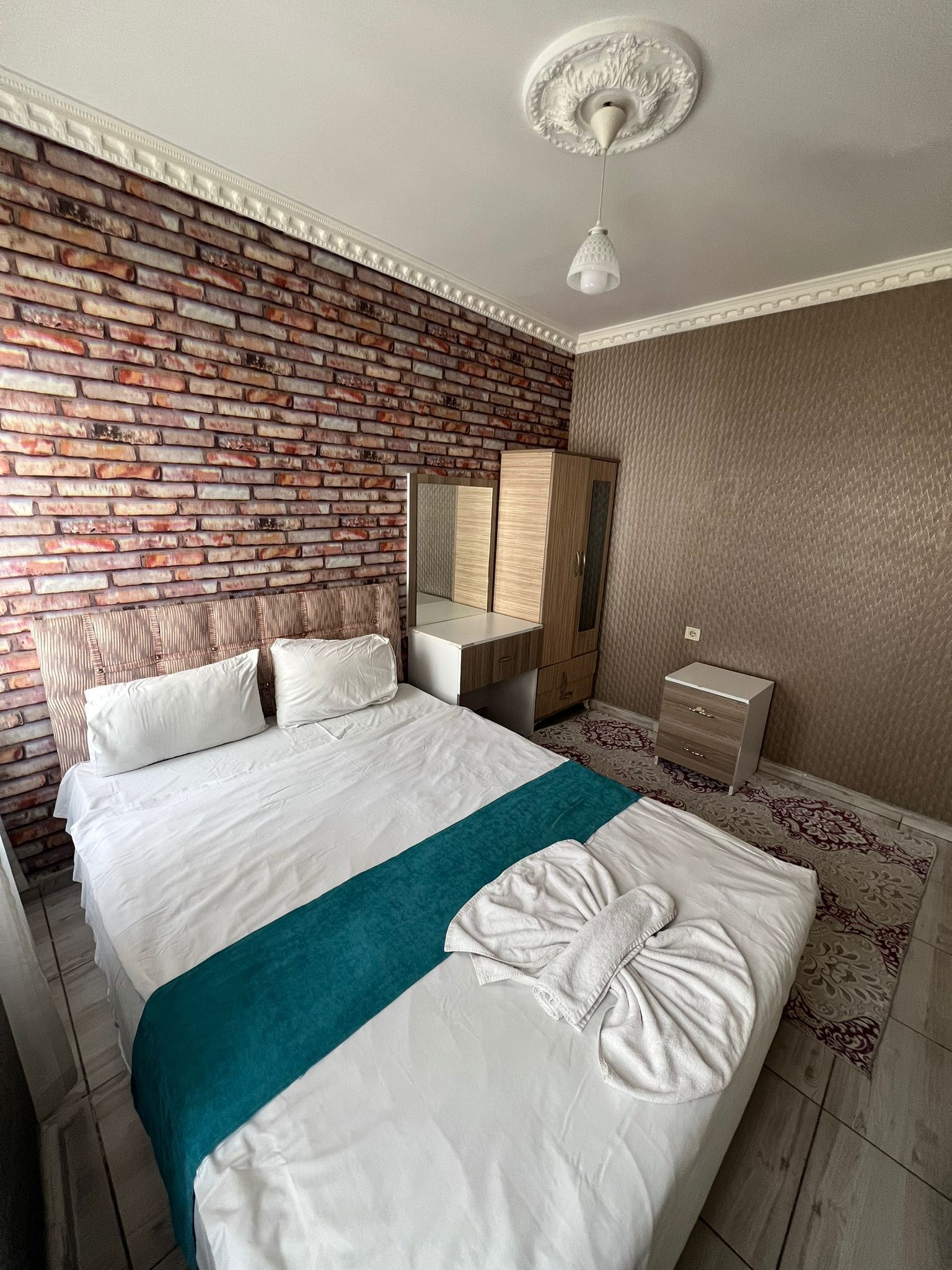 A hotel room with a brick-patterned wall, a bed with white bedding and a teal throw, a wooden nightstand, a mirror, and a hanging light fixture.