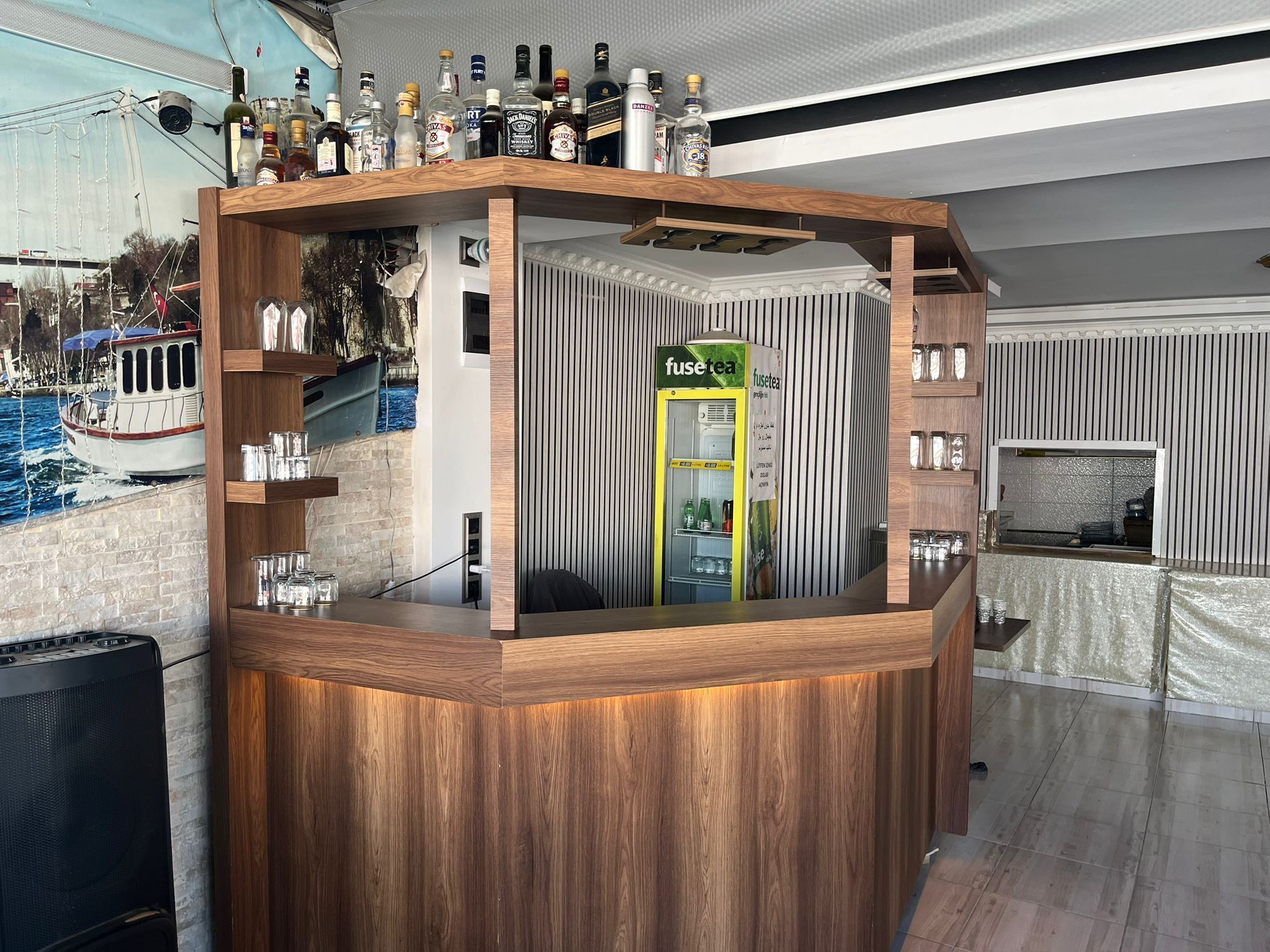 Un espace bar avec un comptoir en bois, un réfrigérateur contenant des bouteilles de « thé infusé » et diverses autres bouteilles.