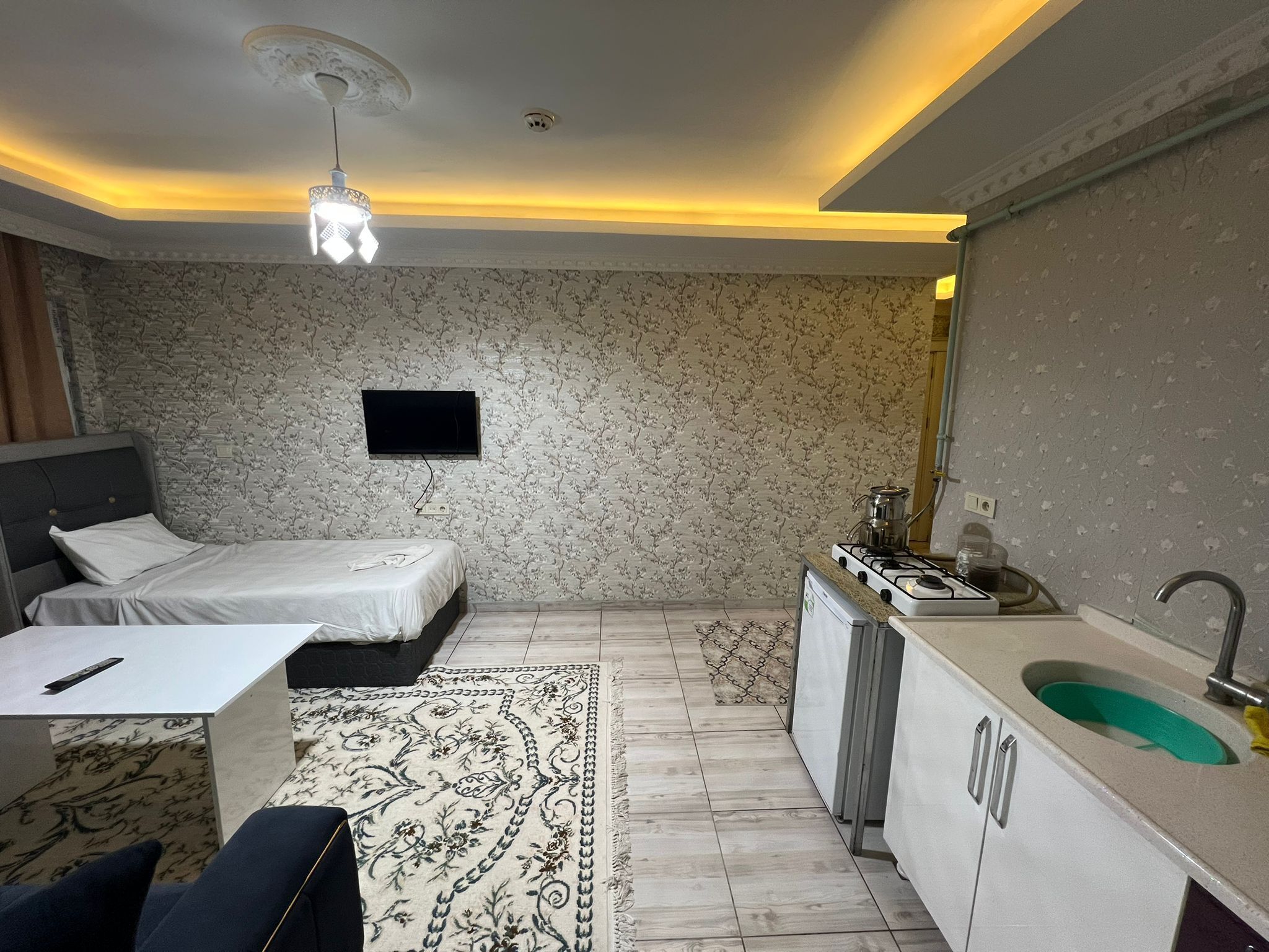 Une chambre avec un lit, une table de chevet et un téléviseur. Il y a une petite kitchenette équipée d’un évier et d’une cuisinière.