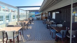 A covered outdoor dining area with tables and chairs, a patterned floor, and a view of buildings through large windows. - Ist Suit Otel (Van)