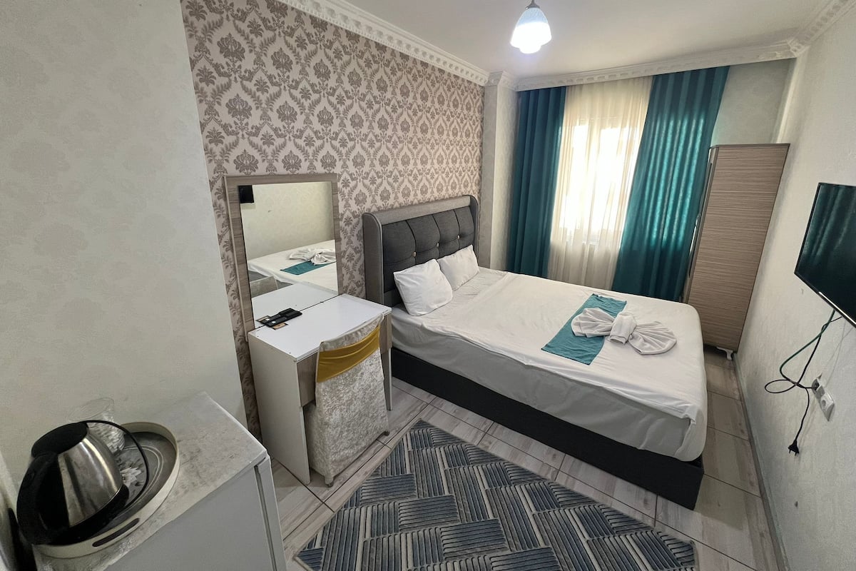 A hotel room with a bed, a bedside table, a mirror, a desk, a chair, a patterned wall, a window with curtains, and a television.