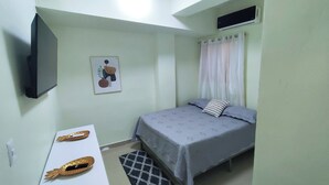 A small, well-lit room with a bed, a flat-screen TV mounted on the wall, a small table with a decorative item, and a window with curtains.