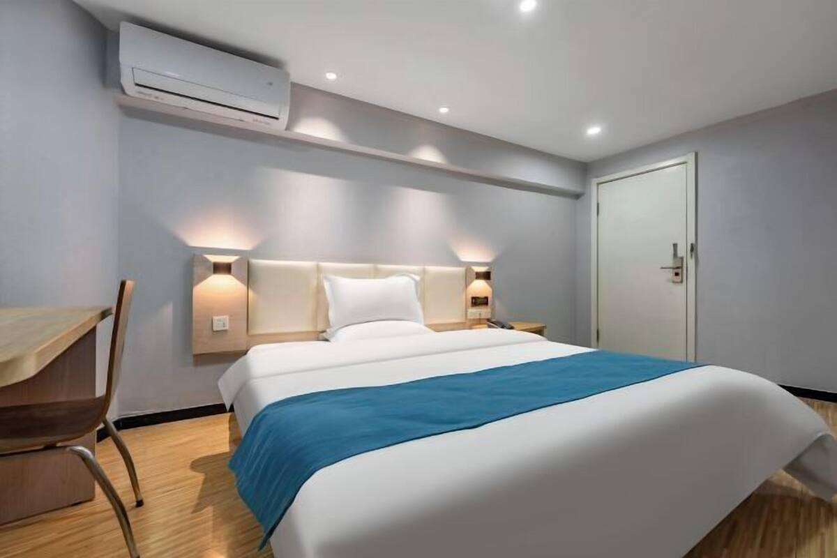 A hotel room with a bed, bedside tables, an air conditioning unit, and a door.