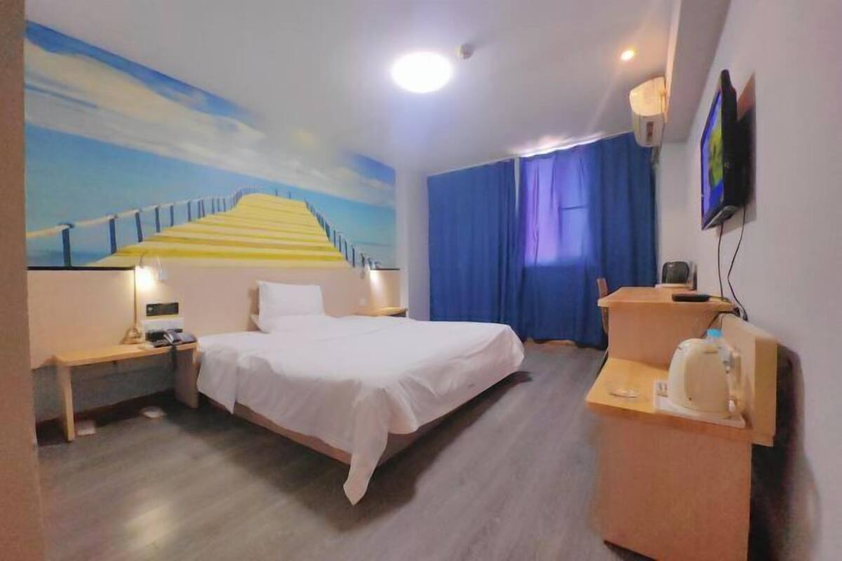 A hotel room with a bed, a desk with a computer, a TV, and a painting of a staircase.