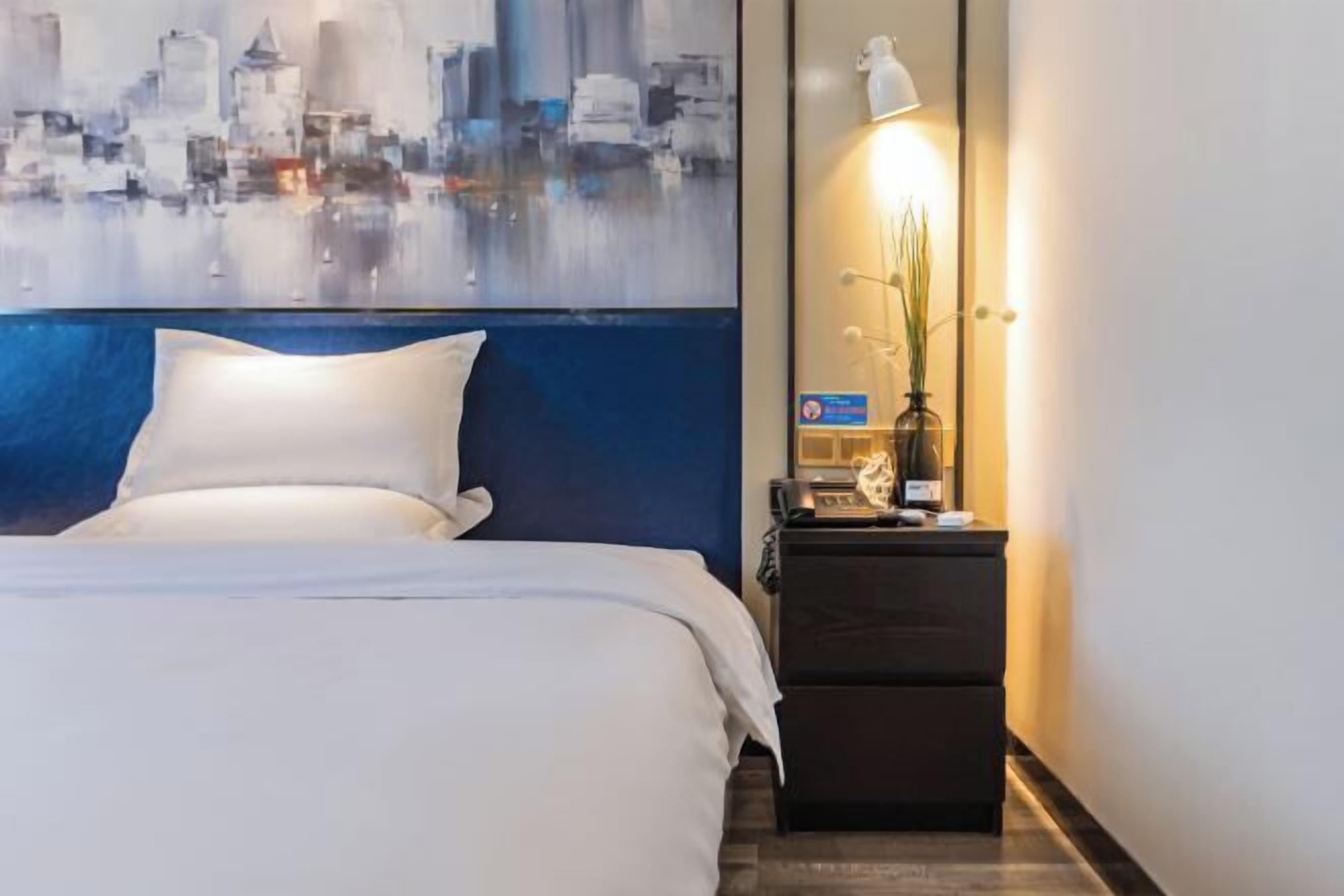 Una habitación de hotel con una cama, una mesita de noche con lámpara y un cuadro de paisaje urbano en la pared.