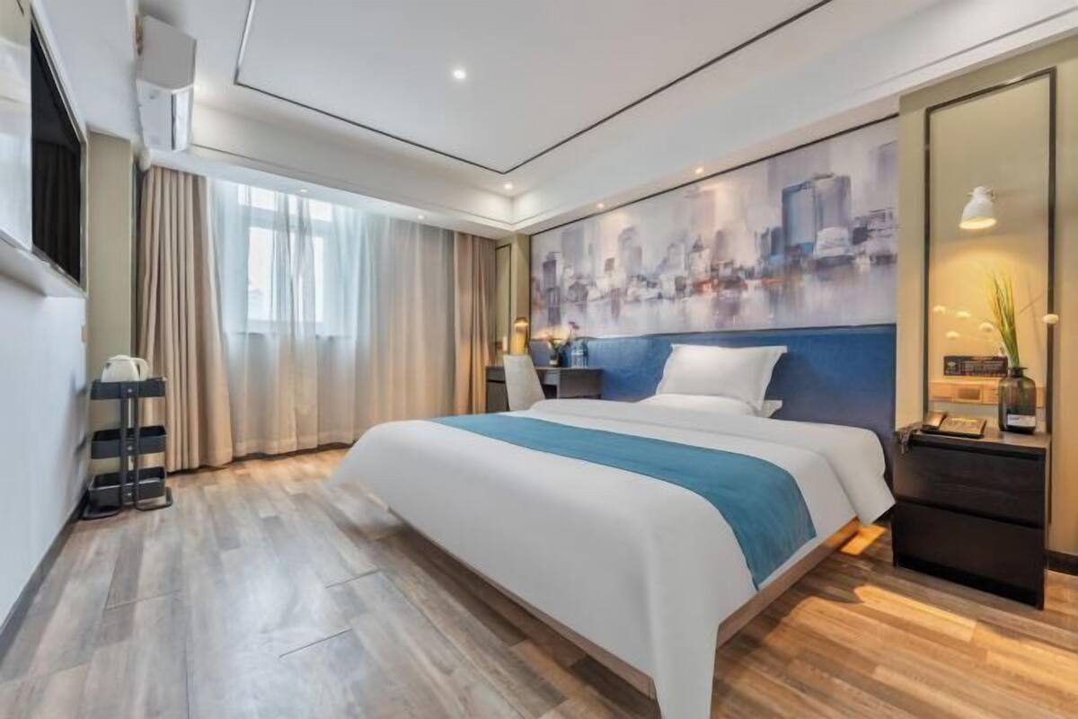 A modern hotel room with a large bed, a cityscape painting on the wall, a nightstand with a lamp, and a wooden floor.
