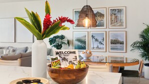 Eine Holzkreisläufer gefüllt mit Früchten und Gemüse, eine weiße Vase mit roten Blumen und ein Schild mit der Aufschrift 'Welcome' auf einem Tisch in einem modernen Wohnzimmer mit einer Couch und gerahmten Bildern an der Wand.