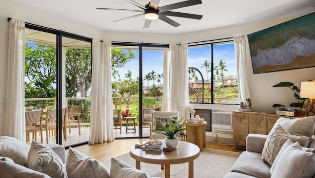 Ein geräumiges Wohnzimmer mit einem großen Schiebeglasfenster, das auf einen Balkon führt, einem Deckenventilator und einem Gemälde mit Strandmotiv.