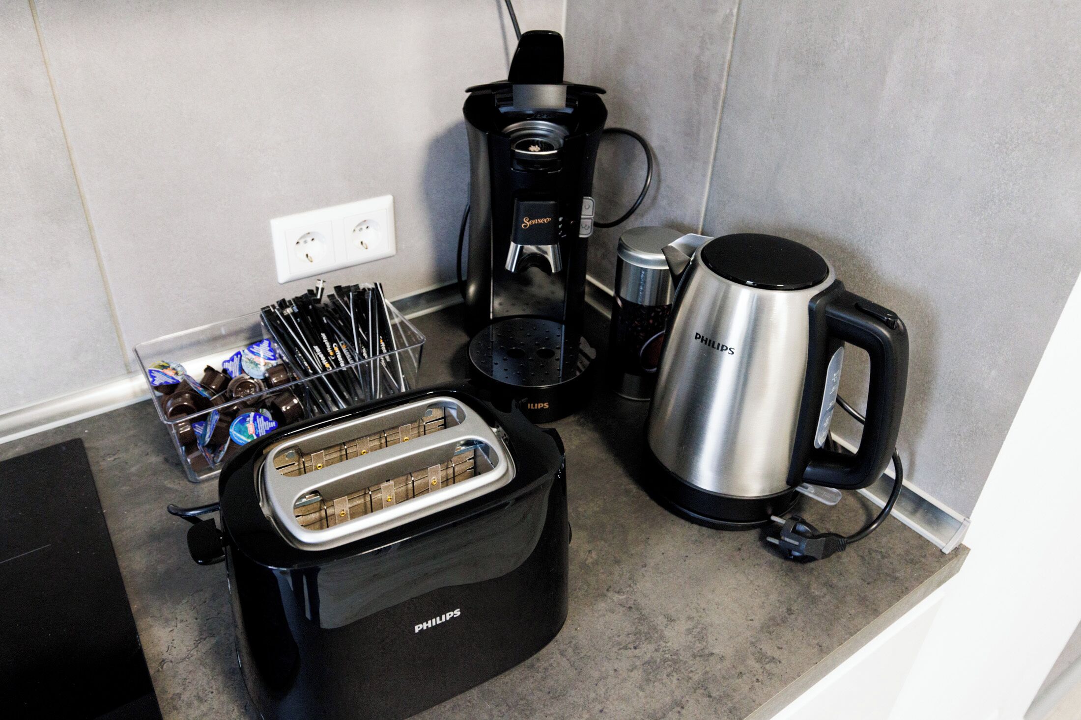 Una cocina con un tostador Philips, una cafetera, un hervidor y un recipiente con cápsulas de café.