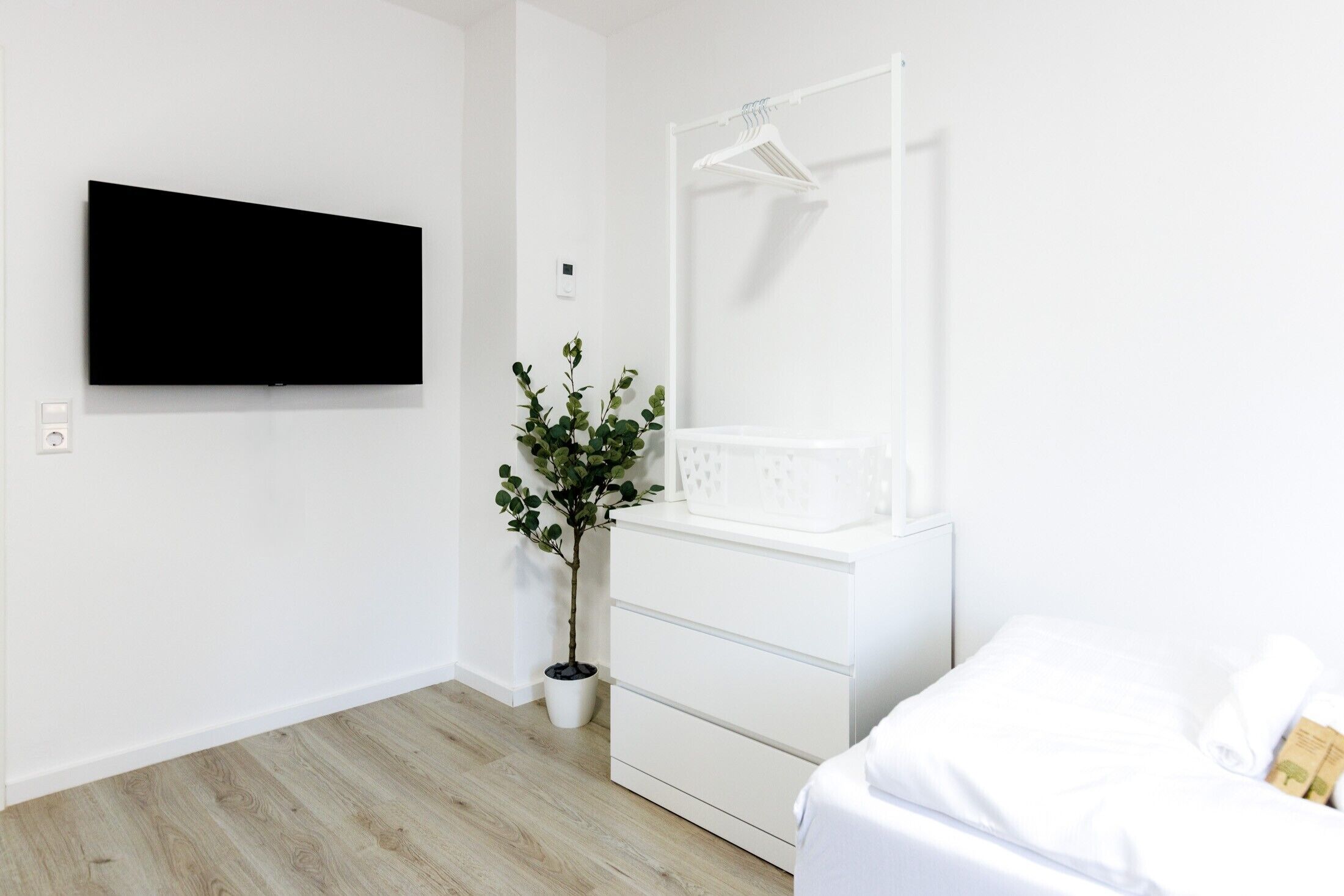 Een moderne, minimalistische slaapkamer met een flatscreen televisie aan de muur, een kleine potplant, een witte dressoir en een bed met witte lakens.