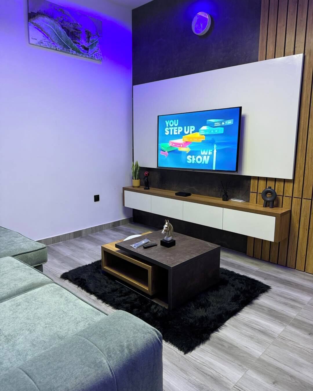 A modern living room with a flat-screen TV mounted on the wall, a wooden cabinet below, a black coffee table, and a grey sofa.