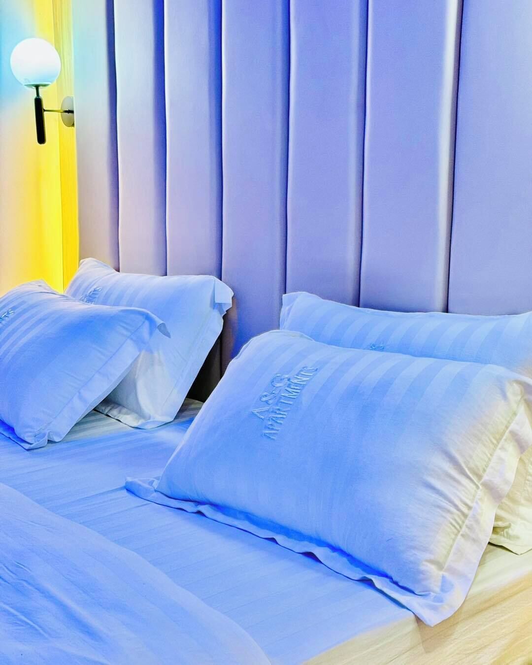 Quarto com cama, travesseiros e luminária fixada na parede.