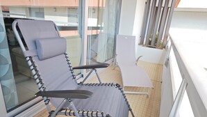 Un balcón con una silla acolchada y una pequeña mesa, ventanas amplias y vistas a edificios.