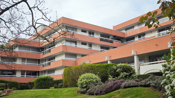 Un edificio residencial de varios pisos con balcones, rodeado de céspedes cuidados y diversos arbustos.