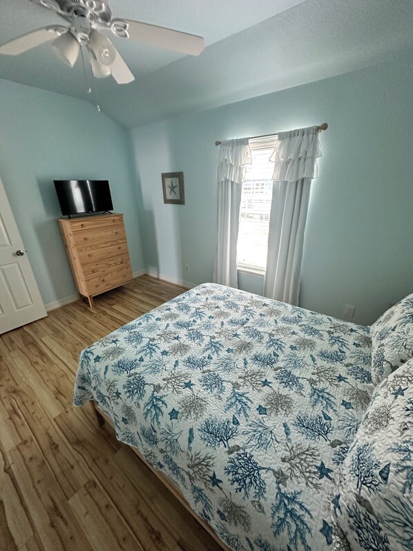 Un dormitorio con cama, cómoda, ventilador de techo, ventana con cortinas e imagen enmarcada en la pared.