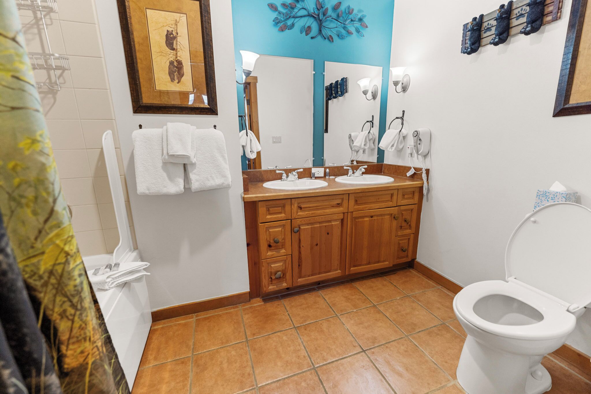 Une salle de bain avec deux lavabos, des toilettes et une douche. Des serviettes sont accrochées au mur et un tableau encadré est suspendu au-dessus des lavabos.