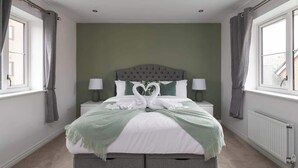 Ein ordentlich eingerichtetes Schlafzimmer mit einem zentral platzierten Bett vor einer grünen Wand, begleitet von zwei Nachttischen mit Lampen, Vorhängen an den Fenstern und einem Heizkörper an der Wand.
