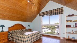 A bedroom with a wooden ceiling, a large window with a plaid valance, a bed with a plaid bedspread, a nightstand with a lamp, and a shelf with decorative items. - Private Pool & Stunning Mountain Views (Tarragona)