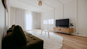 A modern living room with a large flat-screen TV on a wooden stand, a green sofa, a white shag rug, and a large window with sheer curtains.