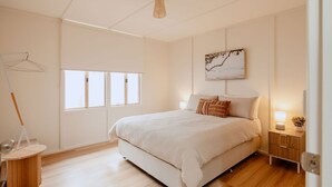 A bedroom with a bed, a bedside table with a lamp, a hanging light fixture, a window, and a wall-mounted artwork.
