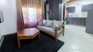 Ein Wohnbereich mit einer Couch, einem Holztisch und einem schwarzen Teppich. Im Hintergrund ist eine Küche mit Schränken und Geräten zu sehen.