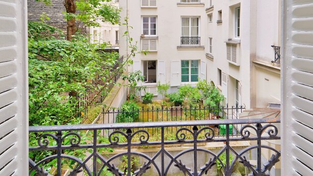 A view from a balcony overlooking a narrow courtyard with a small garden, a building with multiple windows and balconies, and a metal fence.