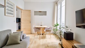 A modern living room with a grey sofa, a wooden dining table with two chairs, a TV mounted on the wall, and a potted plant.