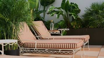 A sunlit outdoor seating area with striped lounge chairs and a small table, surrounded by lush tropical plants.