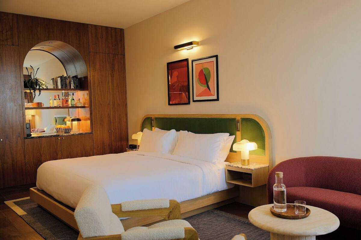 A hotel room with a large bed, a wooden wardrobe, a small table with a bottle, and a red armchair.