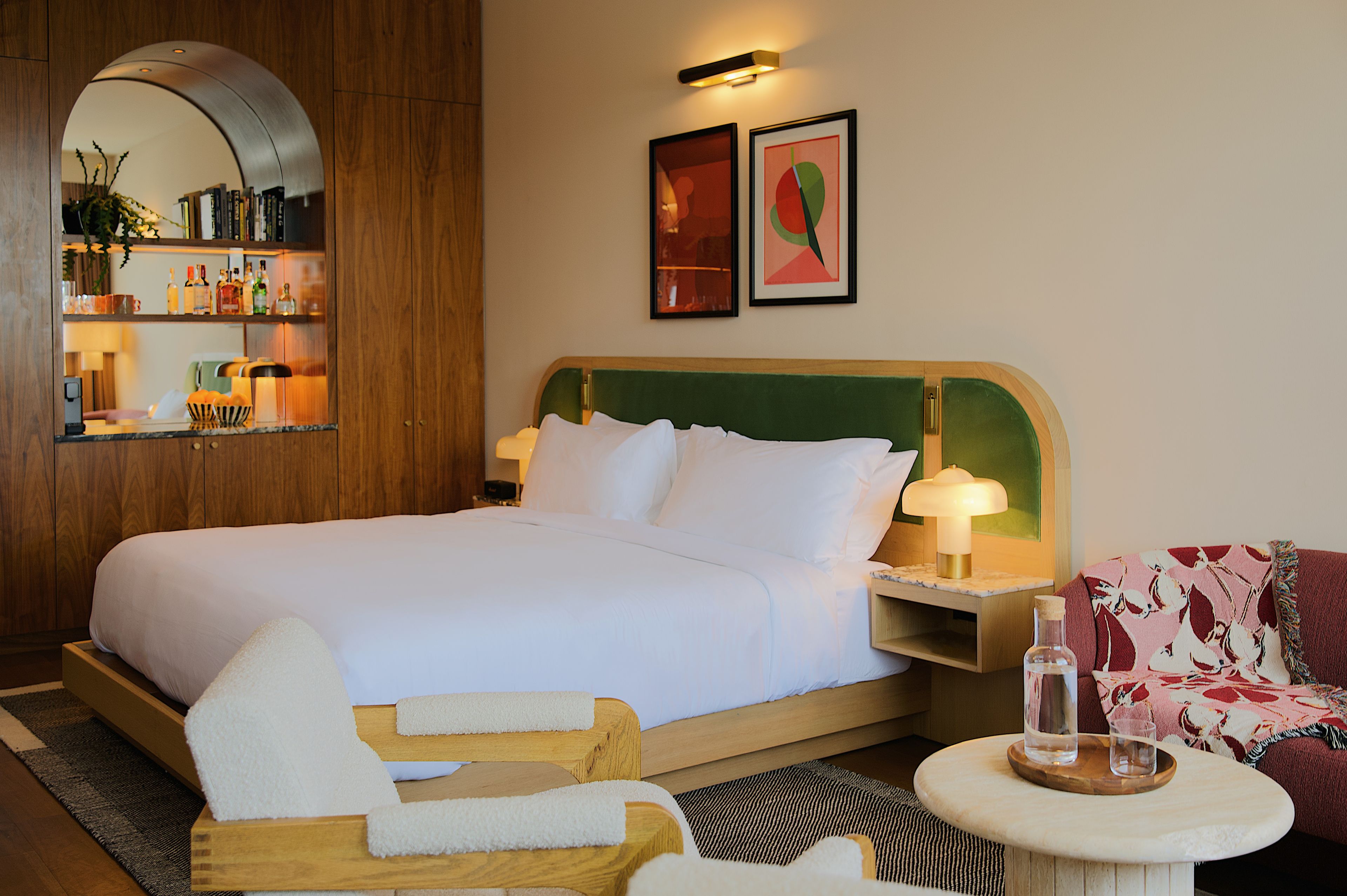 Une chambre d’hôtel avec un grand lit, une tête de lit en bois, une table de chevet avec une lampe, une petite table ronde avec une bouteille et un verre, une chaise et une bibliothèque contenant divers objets.