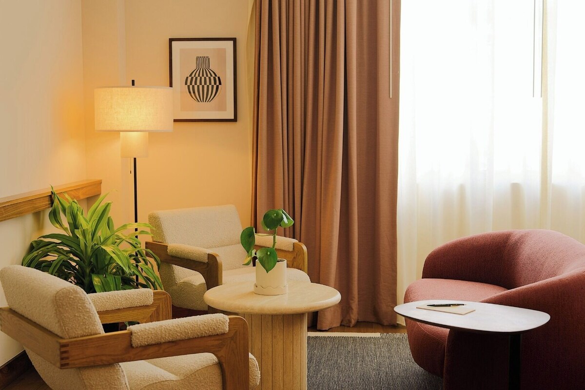 A hotel room with a sofa, armchair, a small round table, a lamp, a potted plant, and a window with curtains.
