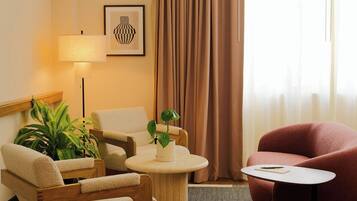 A hotel room with a sofa, armchair, a small round table, a lamp, a potted plant, and a window with curtains.