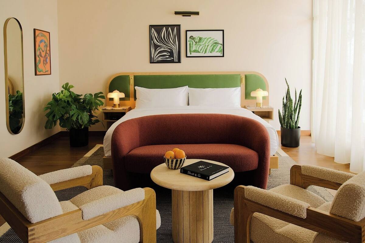 A hotel room with a bed, two bedside tables, a round coffee table, two armchairs, and two potted plants.