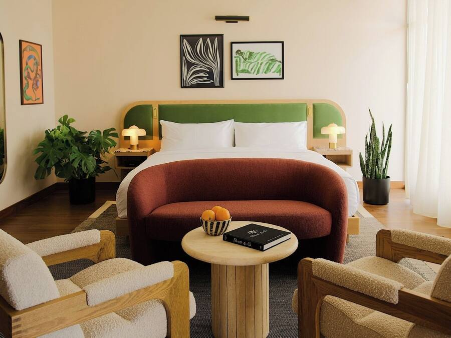 Quarto de hotel com cama, duas mesas de cabeceira, uma mesa de centro redonda, duas poltronas e duas plantas em vasos.