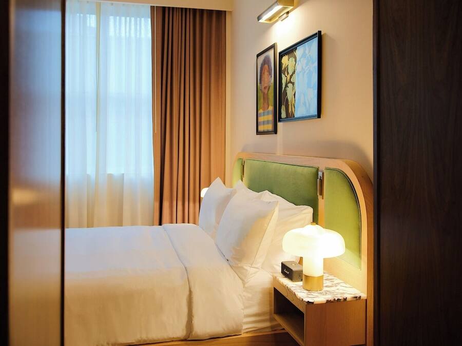 A hotel room with a bed, a nightstand with a lamp, a window with curtains, and two framed artworks on the wall.
