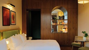 Een hotelkamer met een groot bed, een klein tafeltje met een stoel, een spiegel en een plank met boeken en flessen.