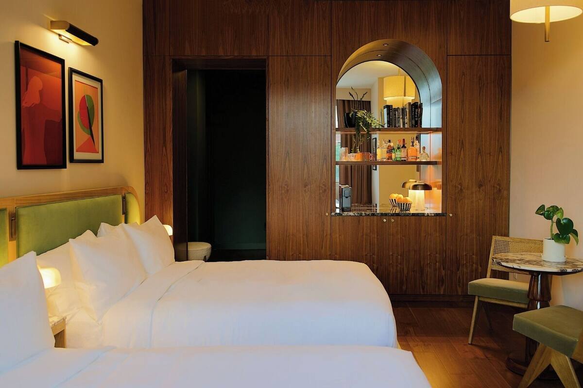 A hotel room with a large bed, a small table with a chair, a mirror, and a shelf with books and bottles.