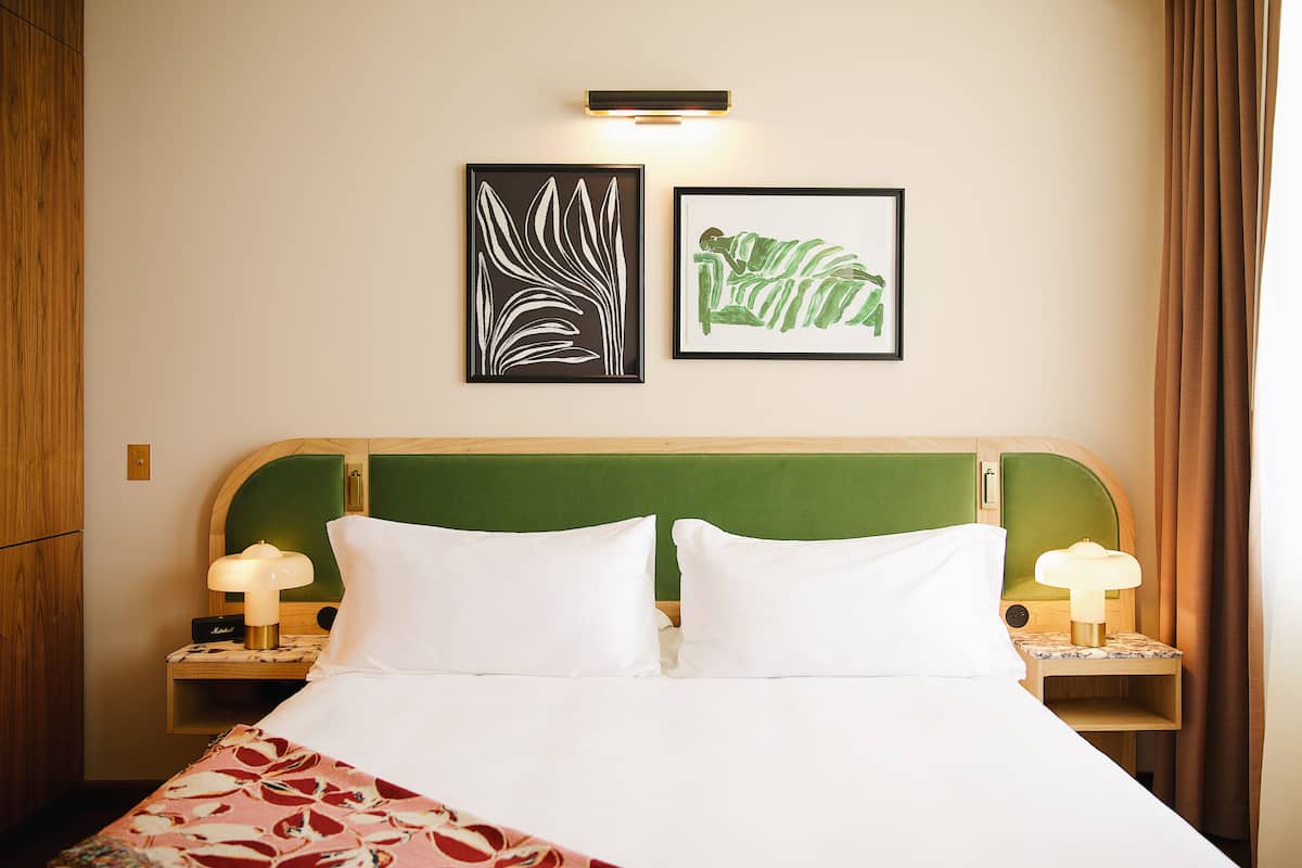 A hotel room with a bed, two bedside tables with lamps, a green headboard, and two framed artworks on the wall.