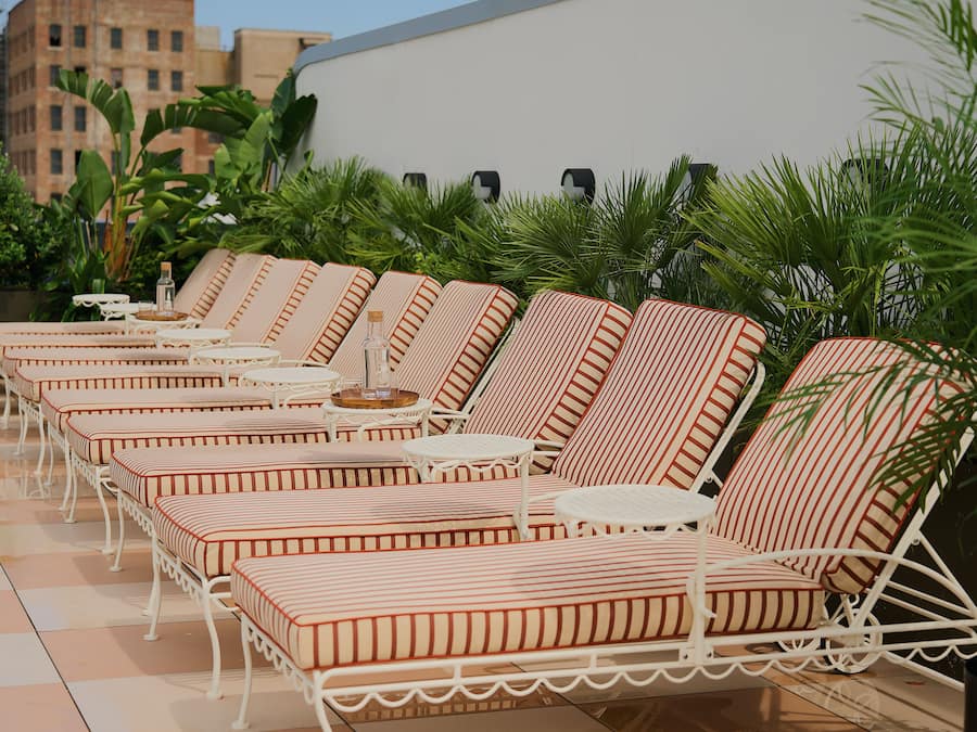 A row of striped lounge chairs with white metal frames and cushions, arranged on a tiled patio with palm plants in the background.