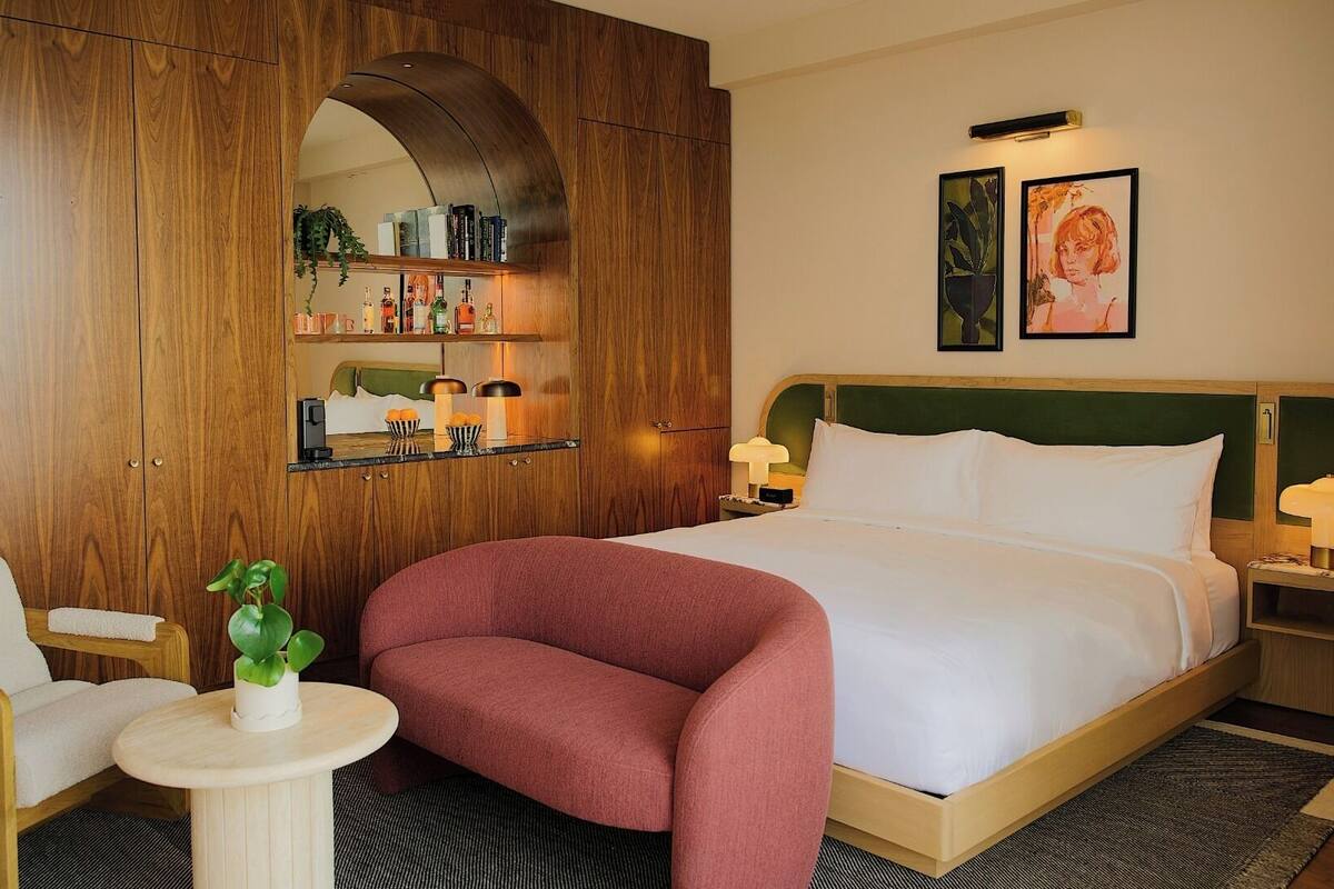 A hotel room with a bed, a wooden wardrobe, a small round table with a plant, a chair, and a nightstand with a lamp.