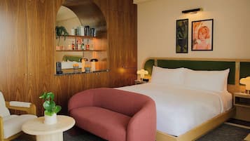 A hotel room with a bed, a wooden wardrobe, a small round table with a plant, a chair, and a nightstand with a lamp.