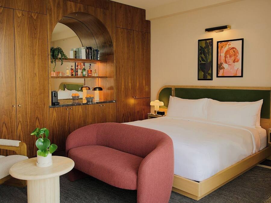 A hotel room with a bed, a wooden wardrobe, a small round table with a plant, a chair, and a nightstand with a lamp.