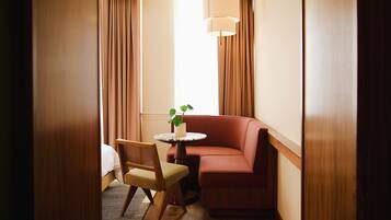 A cozy hotel room with a wooden floor, a red sofa, a small round table with a plant, and a chair.
